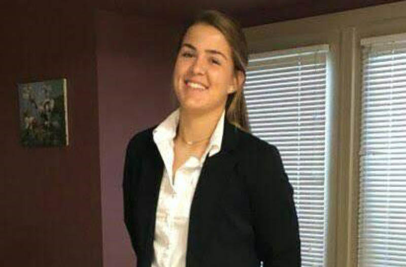 Smiling woman in a dark blazer and white shirt standing indoors with hands behind her back.